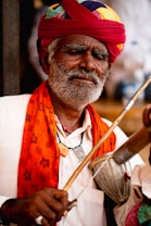 An elderly man with a grey beard and mustache is wearing a colorful turban, white shirt, and an orange shawl. He is playing a traditional stringed instrument and appears to be deeply absorbed in the music.
