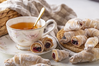 A delicate porcelain teacup filled with amber-colored tea is accompanied by a golden spoon. Surrounding the cup are homemade crescent pastries dusted with powdered sugar, some with visible red and dark fruit jam fillings. The setting includes a woven basket and a soft, neutral-colored cloth in the background, giving a cozy and inviting atmosphere.