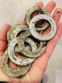 A hand holding several dirty metal washers, covered in soil and debris, on a pale background.