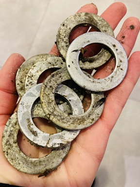 A hand holding several dirty metal washers, covered in soil and debris, on a pale background.