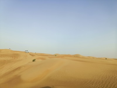 A vast desert landscape with rolling sand dunes extends under a clear, pale blue sky. Two white SUVs navigate the undulating terrain, leaving tire tracks in the sand. Sparse vegetation can be seen scattered across the dunes.