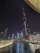 A nighttime cityscape featuring a tall illuminated skyscraper with the words 'THE DUBAI MALL' displayed on it. Surrounding buildings are adorned with bright lights, reflecting off a large body of water in the foreground. A prominent water fountain is visible, adding a dynamic element to the scene.