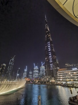 A nighttime cityscape featuring a tall illuminated skyscraper with the words 'THE DUBAI MALL' displayed on it. Surrounding buildings are adorned with bright lights, reflecting off a large body of water in the foreground. A prominent water fountain is visible, adding a dynamic element to the scene.