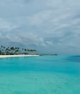 A serene tropical beach scene with calm turquoise waters stretching out under a partly cloudy sky. A small island is lined with tall palm trees on the left, while a series of overwater bungalows extends into the water.