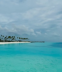 A serene tropical beach scene with calm turquoise waters stretching out under a partly cloudy sky. A small island is lined with tall palm trees on the left, while a series of overwater bungalows extends into the water.