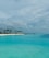 A serene tropical beach scene with calm turquoise waters stretching out under a partly cloudy sky. A small island is lined with tall palm trees on the left, while a series of overwater bungalows extends into the water.