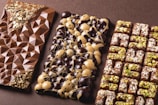 Three artisanal chocolate bars are arranged side by side. The first bar features a geometric pattern with gold flakes embedded at the top and bottom corners. The middle bar is decorated with a mix of dark and white chocolate circular domes. The third bar has a grid pattern, topped with chopped nuts and possibly pistachio pieces.