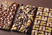 Three artisanal chocolate bars are arranged side by side. The first bar features a geometric pattern with gold flakes embedded at the top and bottom corners. The middle bar is decorated with a mix of dark and white chocolate circular domes. The third bar has a grid pattern, topped with chopped nuts and possibly pistachio pieces.