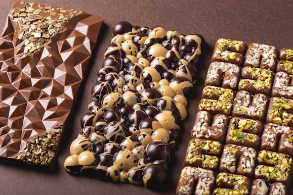 Three artisanal chocolate bars are arranged side by side. The first bar features a geometric pattern with gold flakes embedded at the top and bottom corners. The middle bar is decorated with a mix of dark and white chocolate circular domes. The third bar has a grid pattern, topped with chopped nuts and possibly pistachio pieces.