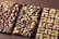 Three artisanal chocolate bars are arranged side by side. The first bar features a geometric pattern with gold flakes embedded at the top and bottom corners. The middle bar is decorated with a mix of dark and white chocolate circular domes. The third bar has a grid pattern, topped with chopped nuts and possibly pistachio pieces.