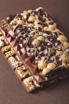 A gourmet chocolate bar is adorned with a variety of toppings. The bar features a rich, dark chocolate base with geometric patterns. On top, there are glossy chocolate orbs swirled with caramel hues, adding a luxurious and tempting appearance. The sides of the bar are embellished with nuts, possibly pistachios and almonds, adding texture and color contrast.