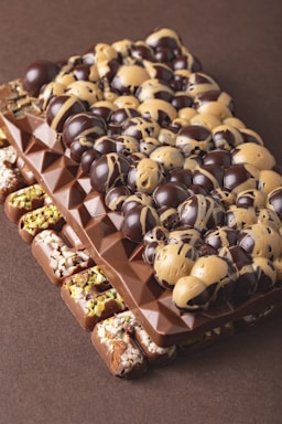 A gourmet chocolate bar is adorned with a variety of toppings. The bar features a rich, dark chocolate base with geometric patterns. On top, there are glossy chocolate orbs swirled with caramel hues, adding a luxurious and tempting appearance. The sides of the bar are embellished with nuts, possibly pistachios and almonds, adding texture and color contrast.