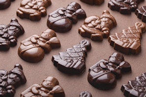 Various chocolate sculptures are shaped into festive holiday figures, including snowmen, Christmas trees, and Santas. The chocolates display a glossy surface with different shades of brown, arranged neatly on a flat surface.