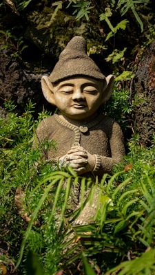 A small stone statue of a figure with elf-like ears and a pointed hat sits among lush green foliage. Its hands are clasped together, and it has a peaceful expression on its face. The setting is natural, with a backdrop of moss-covered rocks and various plants surrounding the statue.