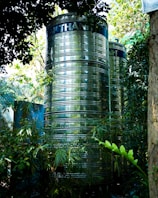Two large, cylindrical, metallic water tanks stand amidst lush, green foliage. The tanks have a shiny, reflective surface and are partially obscured by leaves and branches. Sunlight filters through the canopy above, casting dappled light and shadows on the scene.