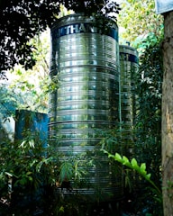 Two large, cylindrical, metallic water tanks stand amidst lush, green foliage. The tanks have a shiny, reflective surface and are partially obscured by leaves and branches. Sunlight filters through the canopy above, casting dappled light and shadows on the scene.