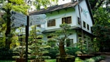 A well-maintained garden with various bonsai trees is in front of a two-story white building. The building has multiple windows, and its roof is slanted with dark shingles. To the left, there is a signpost with multiple directional signs indicating different animals and attractions, including elephants, hippos, and flamingos. The surrounding area is lush with greenery and trees.