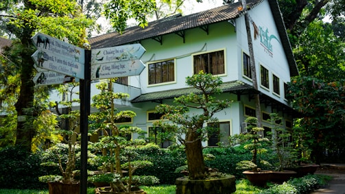 A well-maintained garden with various bonsai trees is in front of a two-story white building. The building has multiple windows, and its roof is slanted with dark shingles. To the left, there is a signpost with multiple directional signs indicating different animals and attractions, including elephants, hippos, and flamingos. The surrounding area is lush with greenery and trees.