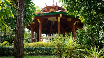 A wooden gazebo with a red-brown structure and green roof is surrounded by lush tropical plants and trees. The gazebo is elevated, with a pathway leading up to it. Bright sunlight filters through the tree leaves, casting dappled shadows on the grass.
