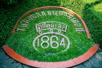 An arrangement of lush green foliage forms the background for white text reading 'THÀO CẦM VIÊN SÀI GÒN', surrounded by a circular pattern of red bricks. At the center, 'THÀNH LẬP 1864' is prominently displayed.