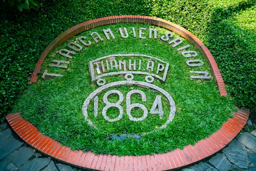 An arrangement of lush green foliage forms the background for white text reading 'THÀO CẦM VIÊN SÀI GÒN', surrounded by a circular pattern of red bricks. At the center, 'THÀNH LẬP 1864' is prominently displayed.