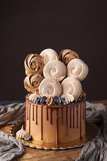 A beautifully decorated cake with rich chocolate layers, topped with artful meringue swirls resembling roses. Accented with fresh blueberries and small silver beads, the cake features a glossy chocolate drip down its sides. It sits on a wooden surface with a dark backdrop, enhancing its elegant aesthetic.