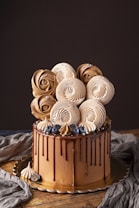 A beautifully decorated cake with rich chocolate layers, topped with artful meringue swirls resembling roses. Accented with fresh blueberries and small silver beads, the cake features a glossy chocolate drip down its sides. It sits on a wooden surface with a dark backdrop, enhancing its elegant aesthetic.
