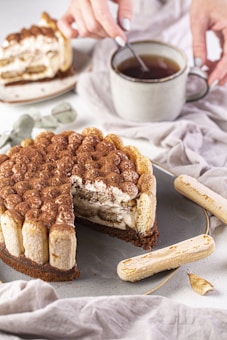 A dessert designed like a cake with layers of creamy filling and topped with cocoa powder is presented on a plate. Nearby, a person's hand stirs a cup of tea, creating a harmonious setting with pieces of ladyfinger biscuits placed around the plate. The atmosphere is accentuated by neutral-colored linens and some green leaves, emphasizing a cozy and inviting mood.