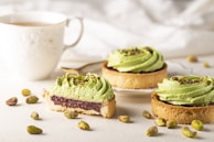 Delicate pistachio tarts are adorned with vibrant green swirls of cream, placed on a light-colored surface. A slice reveals layers of filling, and whole pistachios are scattered around. A white cup with intricate designs holds a hot beverage in the background.