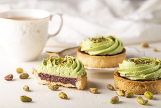 Delicate pistachio tarts are adorned with vibrant green swirls of cream, placed on a light-colored surface. A slice reveals layers of filling, and whole pistachios are scattered around. A white cup with intricate designs holds a hot beverage in the background.