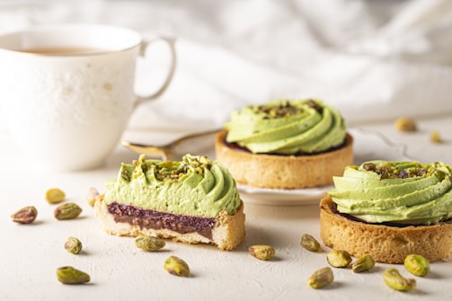 A warm, inviting setting showing a cup of pistachio coffee next to scattered green Antep pistachios on a softly lit beige background.