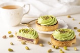 Three tarts with a light green cream topping are arranged on a plate and table, garnished with crushed pistachios. One tart has a bite taken out of it, revealing a layer of jam inside. A cup of tea sits beside the tarts, with a gold-handled spoon resting on the saucer. Pistachios are scattered around on the white surface.