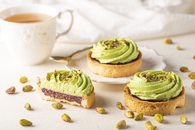 Three tarts with a light green cream topping are arranged on a plate and table, garnished with crushed pistachios. One tart has a bite taken out of it, revealing a layer of jam inside. A cup of tea sits beside the tarts, with a gold-handled spoon resting on the saucer. Pistachios are scattered around on the white surface.