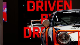 A classic Porsche race car with a bold and colorful paint scheme is prominently displayed. The car's design features vibrant stripes in blue, orange, and white. 'DRIVEN BY DREAMS' is written in large red letters in the background, adding to the dynamic and aspirational atmosphere. A partially visible digital screen on the left side shows an image of a person.
