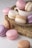 A collection of pastel-colored macarons is arranged elegantly around a white teacup on a woven mat. The macarons come in shades of pink, purple, cream, and brown, with delicate textures and creamy fillings.