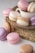 A collection of pastel-colored macarons is arranged elegantly around a white teacup on a woven mat. The macarons come in shades of pink, purple, cream, and brown, with delicate textures and creamy fillings.