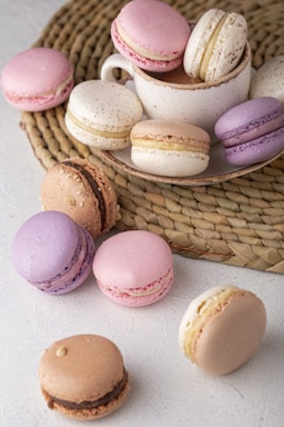 An assortment of colorful macarons is arranged around and inside a beige cup on a textured woven mat. The macarons come in various pastel shades, including pink, purple, and cream, with visible filling in the center. The scene is set on a light textured surface, creating a harmonious presentation of desserts.