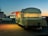 A retro-style food trailer with a rounded top is parked in a lot during twilight. The trailer has a lighted interior visible through large windows adorned with curtains. Its side awning is open, with a warm, inviting glow emanating from inside. A white pickup truck is parked nearby. The background displays a dim skyline, indicating early evening or dawn, with a gradient of blue and orange in the sky.