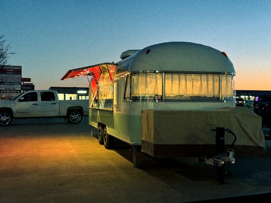 A retro-style food trailer with a rounded top is parked in a lot during twilight. The trailer has a lighted interior visible through large windows adorned with curtains. Its side awning is open, with a warm, inviting glow emanating from inside. A white pickup truck is parked nearby. The background displays a dim skyline, indicating early evening or dawn, with a gradient of blue and orange in the sky.