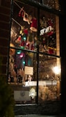 A cozy store window display features festive miniature clothes like red and green Santa and elf outfits hanging on a string with clothespins. Inside, bottles with 'JOY' tags are arranged alongside candles and holiday decorations. Twinkling lights and a warm glow set a seasonal, cheerful atmosphere.