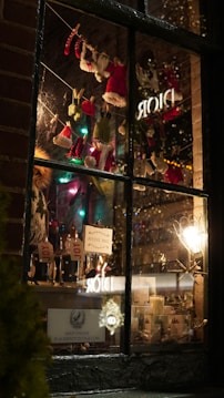 A cozy store window display features festive miniature clothes like red and green Santa and elf outfits hanging on a string with clothespins. Inside, bottles with 'JOY' tags are arranged alongside candles and holiday decorations. Twinkling lights and a warm glow set a seasonal, cheerful atmosphere.