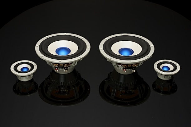 Four speaker subwoofers with blue center domes are arranged symmetrically against a black background, showcasing different sizes from left to right. The speakers have a metallic frame with black rubber surrounds and reflect light subtly, adding depth to the image.