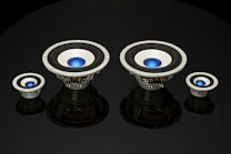 Four speaker subwoofers with blue center domes are arranged symmetrically against a black background, showcasing different sizes from left to right. The speakers have a metallic frame with black rubber surrounds and reflect light subtly, adding depth to the image.