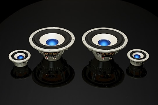 Four speaker subwoofers with blue center domes are arranged symmetrically against a black background, showcasing different sizes from left to right. The speakers have a metallic frame with black rubber surrounds and reflect light subtly, adding depth to the image.