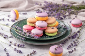 A stack of colorful macarons is artfully arranged on a dark green ceramic plate. Each macaron is topped with small lavender flowers, creating a charming and delicate visual effect. Surrounding the plate are sprigs of fresh lavender, enhancing the aesthetic with a natural and rustic touch. The background is a soft, light-colored fabric, providing a neutral backdrop that allows the vibrant colors of the macarons to stand out.