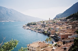 A scenic coastal town nestled between a clear blue lake and towering mountains. The town is characterized by its terracotta rooftops and colorful buildings lining the waterfront. Boats dot the tranquil waters, and lush greenery is visible, creating a picturesque landscape.