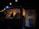 A dimly lit food truck is decorated with hanging string lights. Inside, a person works at the counter, surrounded by menus and cooking equipment. The exterior features a dartboard mounted on a wooden wall, contributing to the cozy, informal atmosphere.
