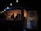 A dimly lit food truck is decorated with hanging string lights. Inside, a person works at the counter, surrounded by menus and cooking equipment. The exterior features a dartboard mounted on a wooden wall, contributing to the cozy, informal atmosphere.