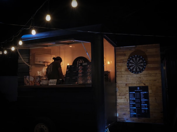 A dimly lit food truck is decorated with hanging string lights. Inside, a person works at the counter, surrounded by menus and cooking equipment. The exterior features a dartboard mounted on a wooden wall, contributing to the cozy, informal atmosphere.