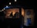 A dimly lit food truck is decorated with hanging string lights. Inside, a person works at the counter, surrounded by menus and cooking equipment. The exterior features a dartboard mounted on a wooden wall, contributing to the cozy, informal atmosphere.